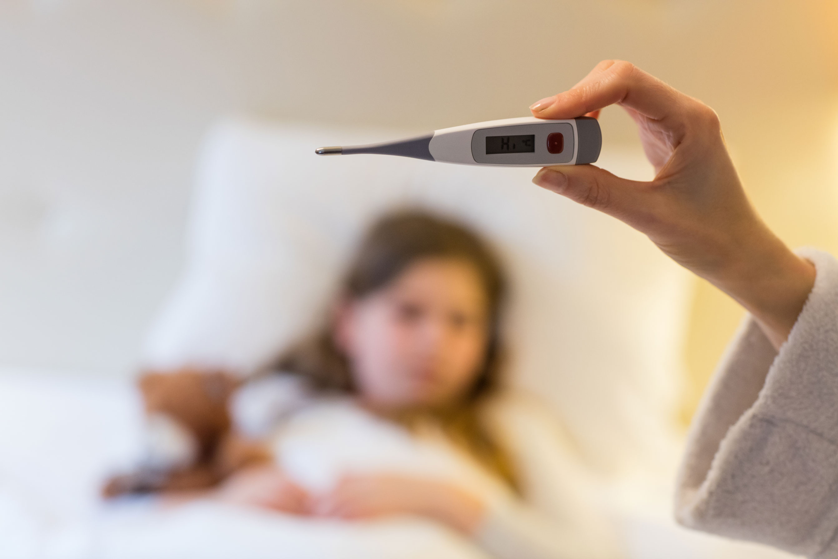 Mão segurando termômetro digital mostrando febre alta com criança doente deitada ao fundo, representando sintomas da gripe K